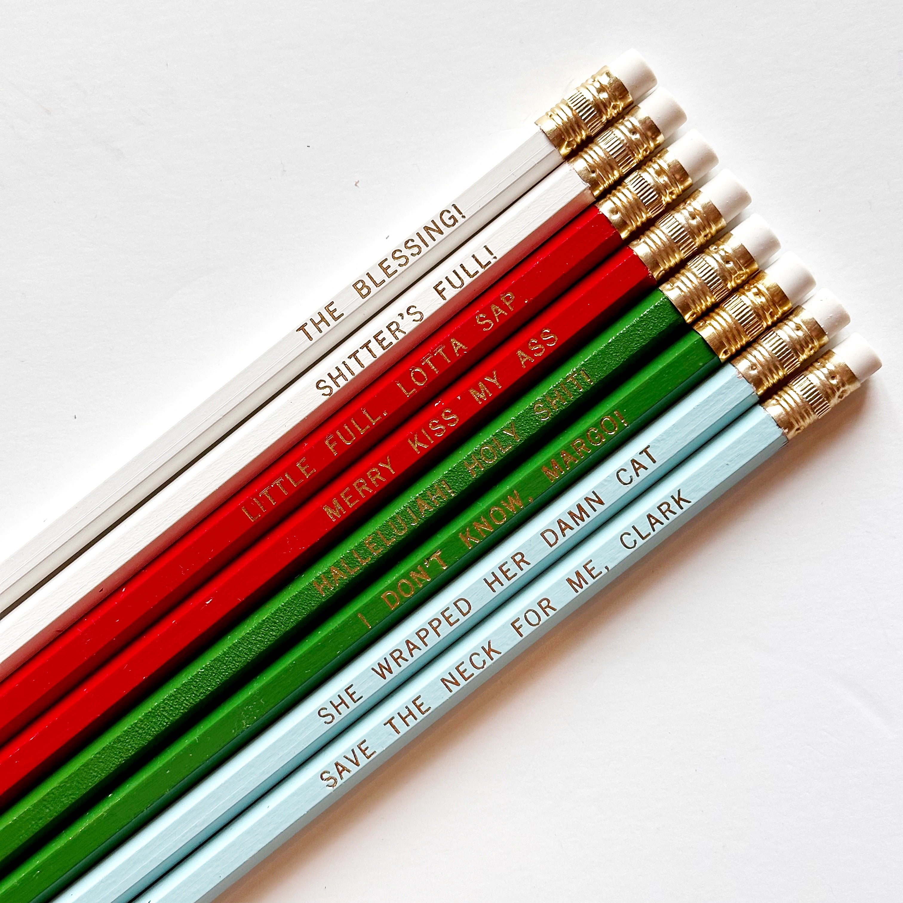 Eight pencils with gold foil text says, "Merry Kiss My Ass Little Full. Lotta Sap. The Blessing! Shitter's Full! Hallelujah! Holy Shit! I Don't Know, Margo! She Wrapped Her Damn Cat Save The Neck For Me, Clark".