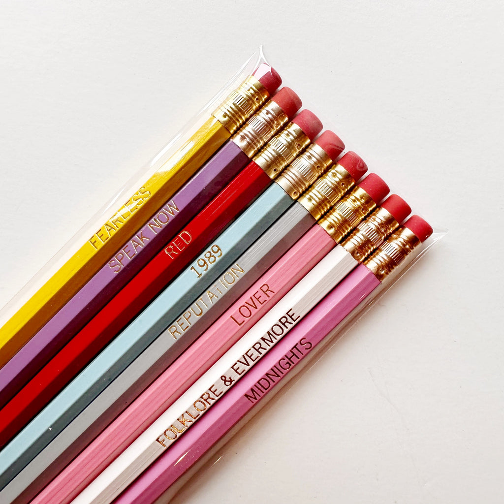 Set of No 2 pencils with Taylor Swift album names in gold text: "Fearless" on yellow pencil, "Speak Now" on lilac, "Red" on red pencil, "1989" on light blue pencil, "Reputation" on grey pencil, "Lover" on pink pencil , "Folklore & Evermore" on white pencil, "Midnights" on dark pink pencil.