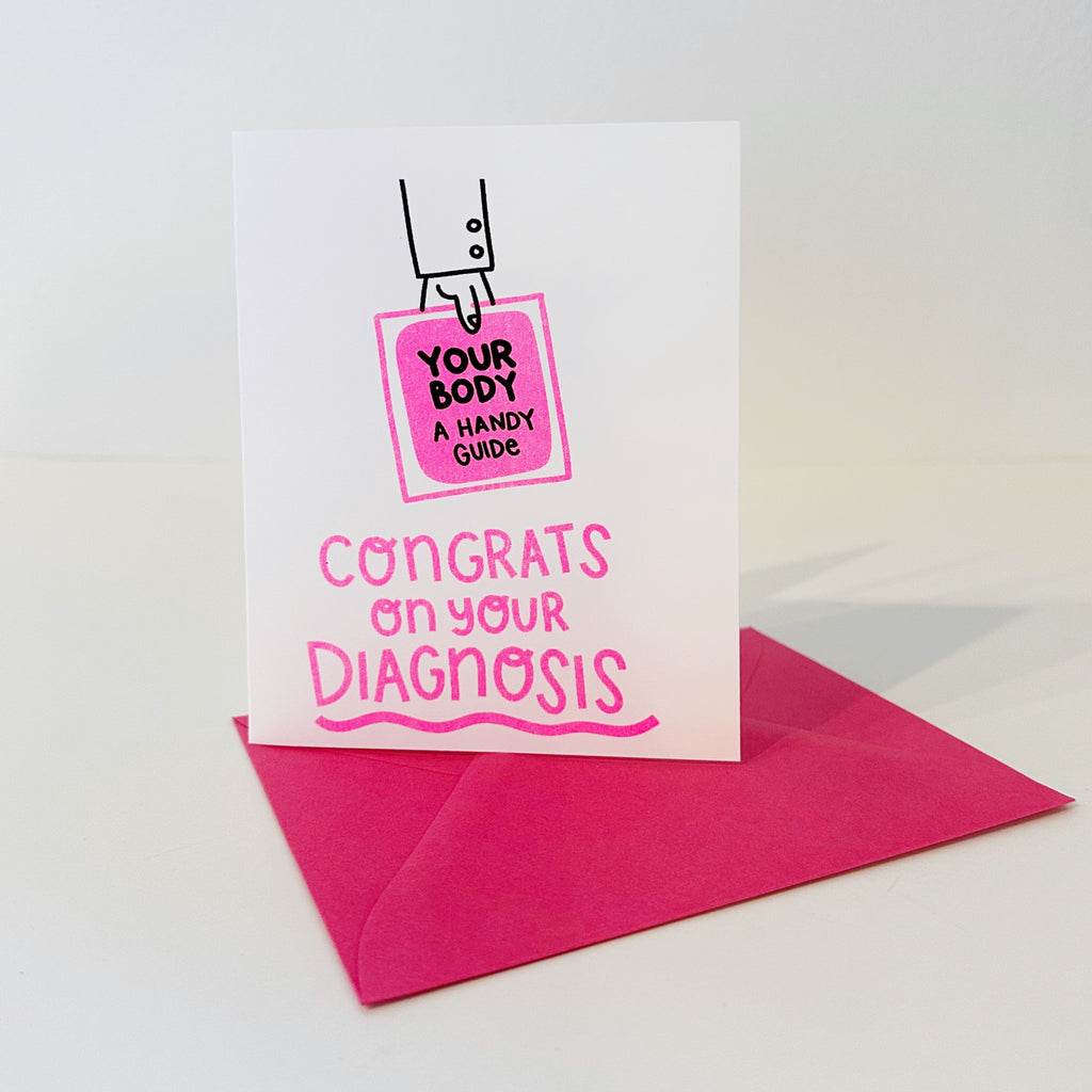 Image of card with white background and image of a hand holing a pink book with black text says, "Your body, a handy guide" and pink text says, "Congrats on your diagnosis". Bright pink envelope included. 