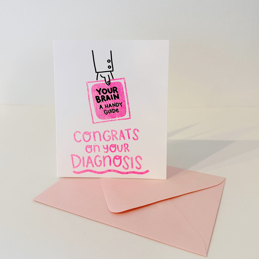 mage of card with white background and image of a hand holing a pink book with black text says, "Your brain, a handy guide" and pink text says, "Congrats on your diagnosis". Bright pink envelope included.