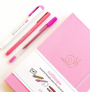 Image of pink journal with image of gold foil snail and pink elastic strap on right side to hold closed with image of three pink pens. 