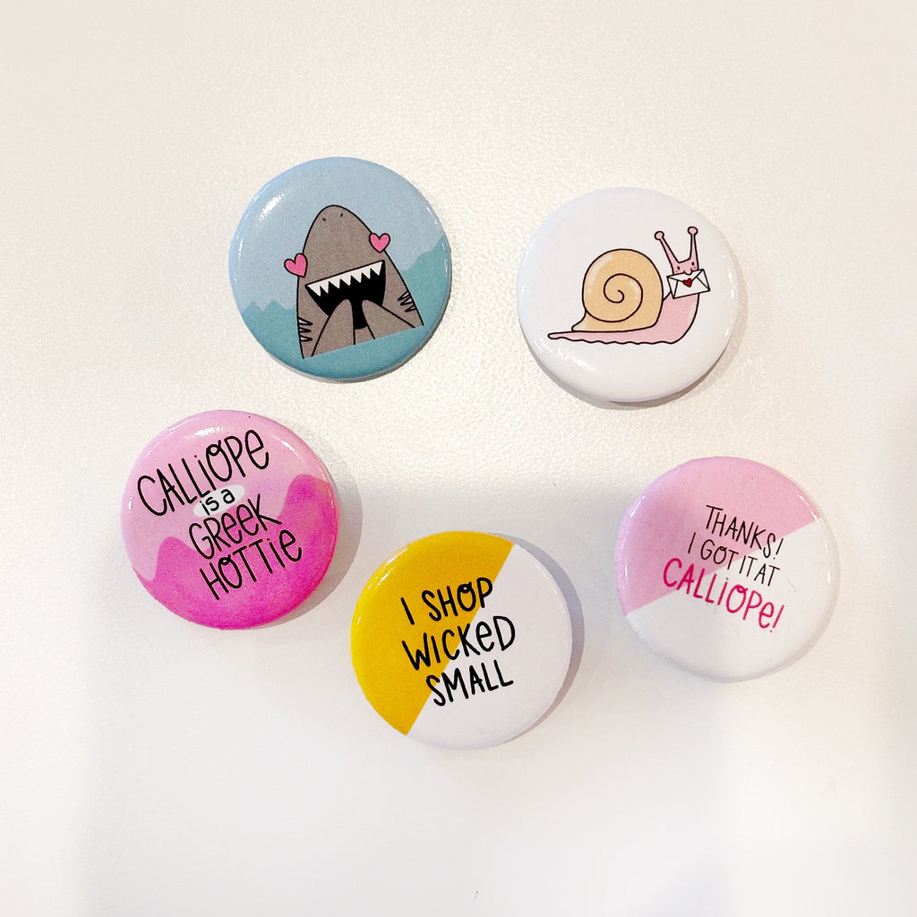 Image of five circular metal buttons. Blue background with image of grey shark with pink heart eyes. White background with image of snail with pink body and yellow shell holding a white letter. Pink and white background with black text says, "Thanks! I got it at" and pink text says, "Calliope". Yellow and white background with black text says, "I shop wicked small". Pink and hot pink background with black text says, "Calliope is a Greek hottie". 