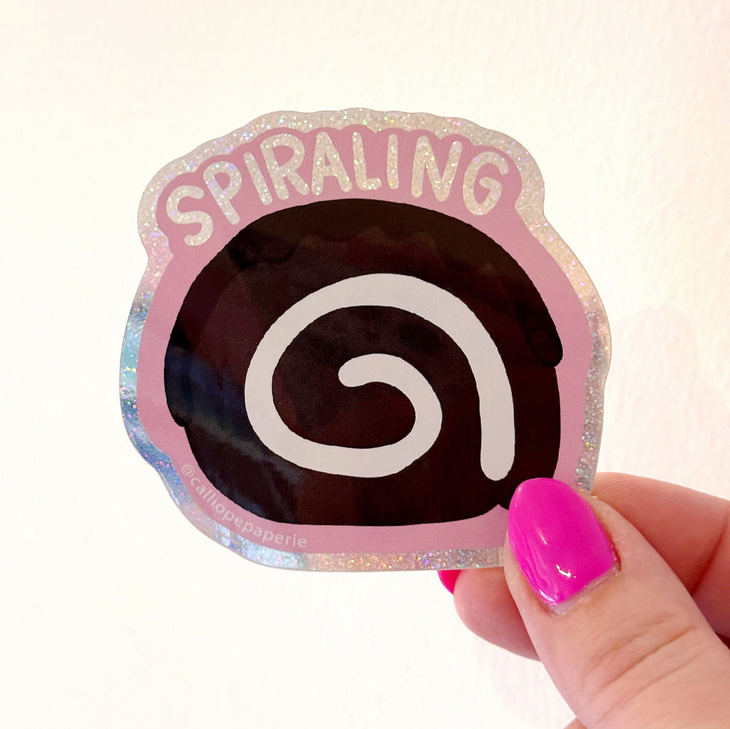 Image of sticker image of a spiral snack cake slice in brown and white with pink and holographic border with holographic text says, "Spiraling". 