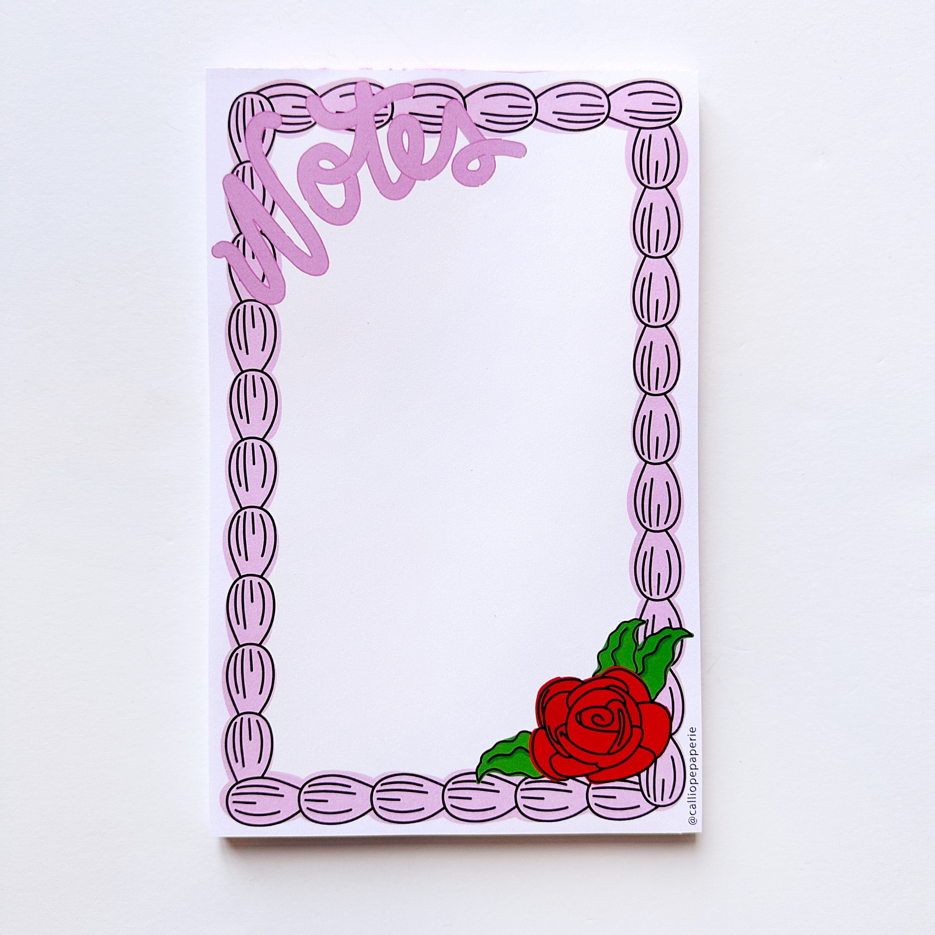 Notepad with white background and pink icing border with pink text says, "Notes" across the top left corner with red rose and green leaves on bottom right corner. 