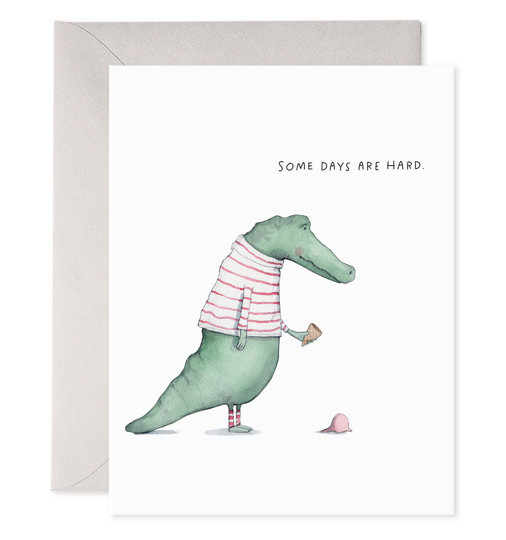 Greeting card with white background  with image of a green alligator wearing a red and white striped shirt holding an ice cream cone with the ice cream on the ground. Black text says, "Some days are hard.". Grey envelope is included. 