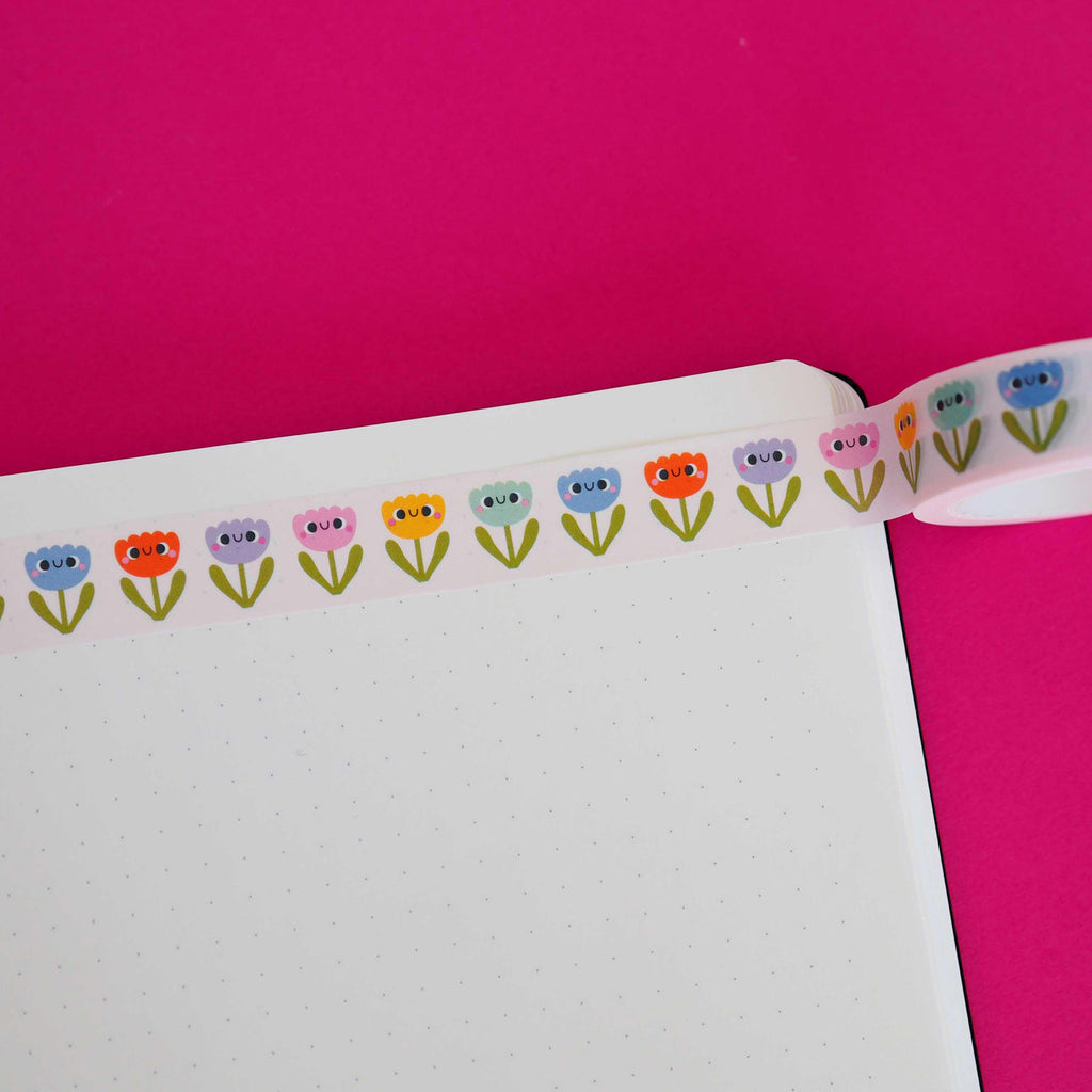 Washi tape with white background and images of flowers with faces in orange, blue, lilac, mint green, and yellow with green stems.