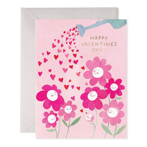 Greeting card with pink background and images of pink and white flowers being watered  with red and pink hearts from a blue watering can. Gold foil text says, "Happy Valentine's Day!". Grey envelope included. 