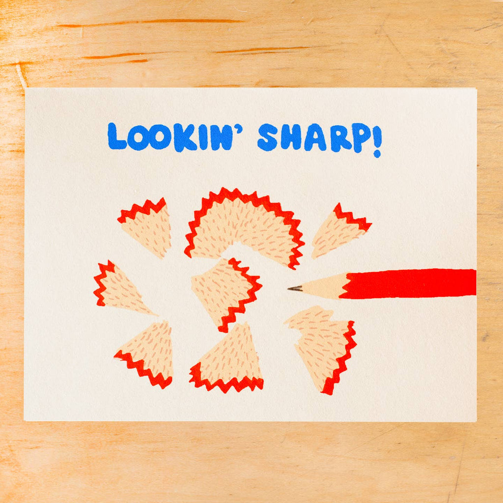 Greeting card with white background with image of a red pencil being sharpened and blue text says, "Lookin' sharp!". Envelope included. 