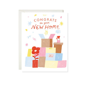 Greeting card with white background and image of brown, pink, blue and yellow boxes with a red potted plant and red text says, "Congrats on your new home". Envelope included. 