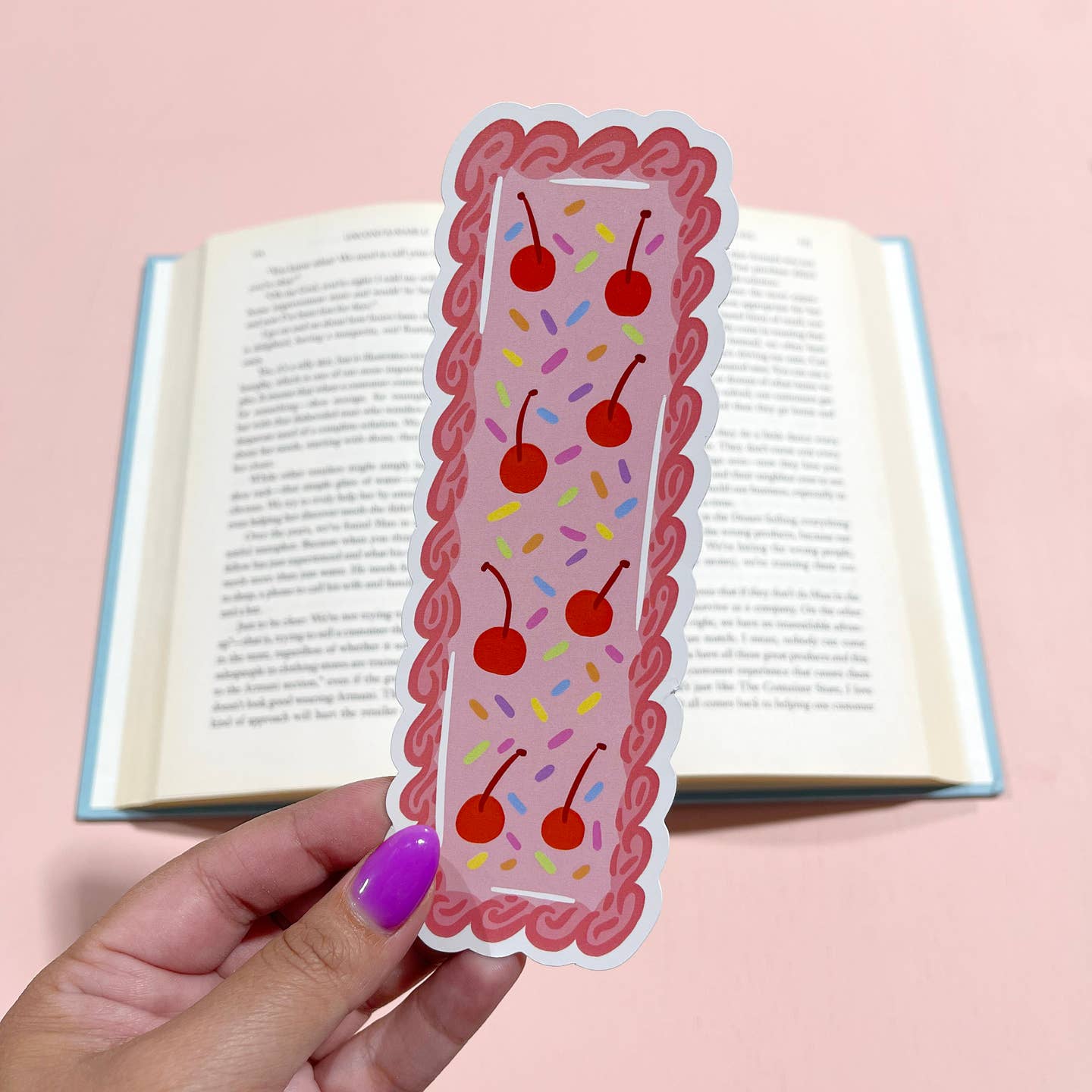 Bookmark in shape of a bar cake with pink background and red frosting outline with sprinkles and red cherries. 