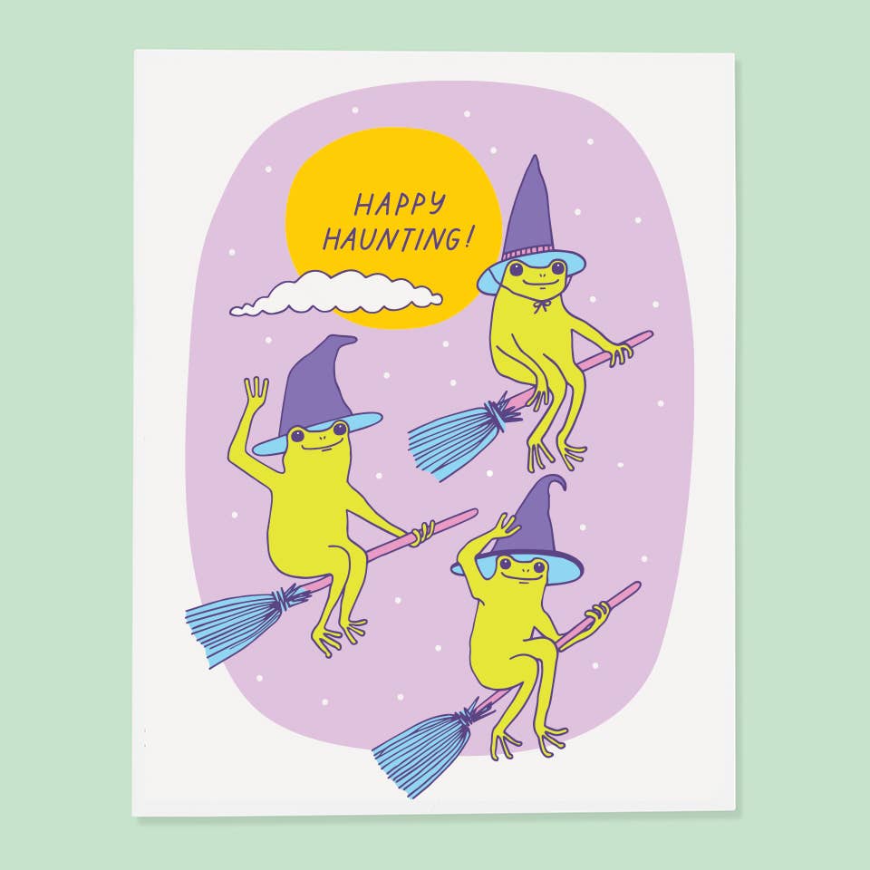 Greeting card with lilac background with white border and images of three green frogs riding brooms and wearing witch hats with yellow moon with purple text says, "Happy Haunting!". Envelope included. 