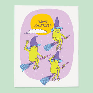 Greeting card with lilac background with white border and images of three green frogs riding brooms and wearing witch hats with yellow moon with purple text says, "Happy Haunting!". Envelope included. 