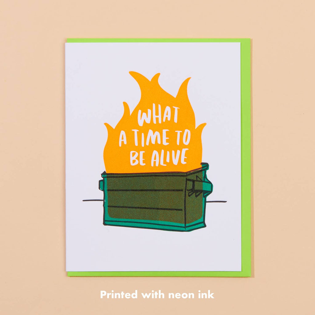 Greeting card with white background and image of a green dumpster with fire and white text says, "What a time to be alive". Green envelope included.