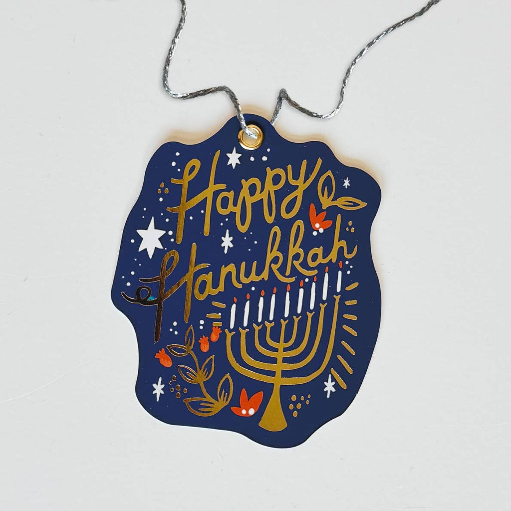 Gift tag with blue background and gold foil text says, "Happy Hanukkah" and image of gold foil menorah with white candles. 