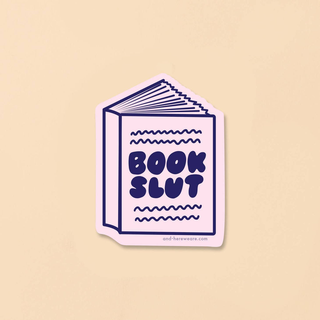 Image of sticker in the shape of a book with  pale pink background and blue text says, "Book slut". 