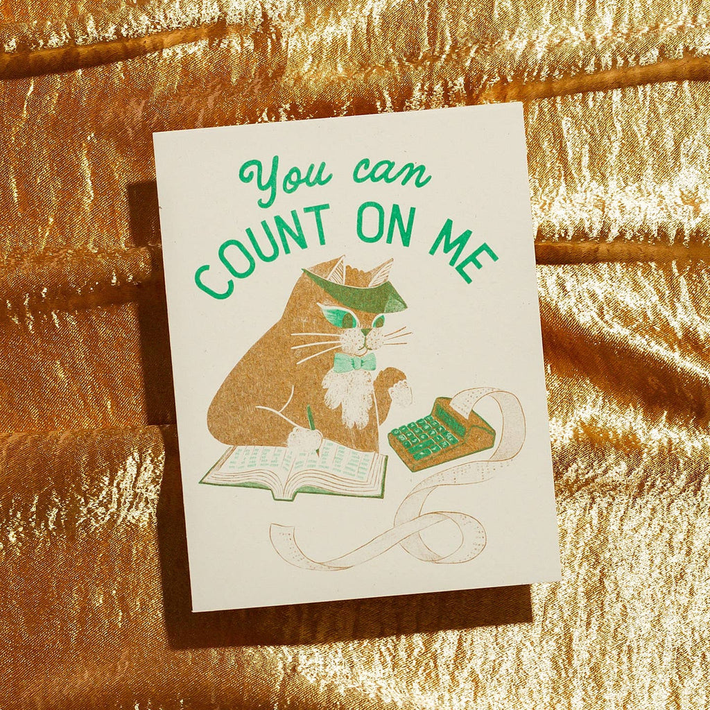 Greeting card with cream background and image of brown cat wearing an accountant visor and using a adding machine and ledger book. Green text says, "You can count on me". Envelope included.