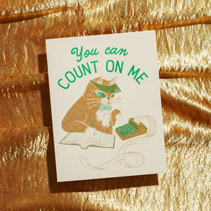 Greeting card with cream background and image of brown cat wearing an accountant visor and using a adding machine and ledger book. Green text says, "You can count on me". Envelope included.