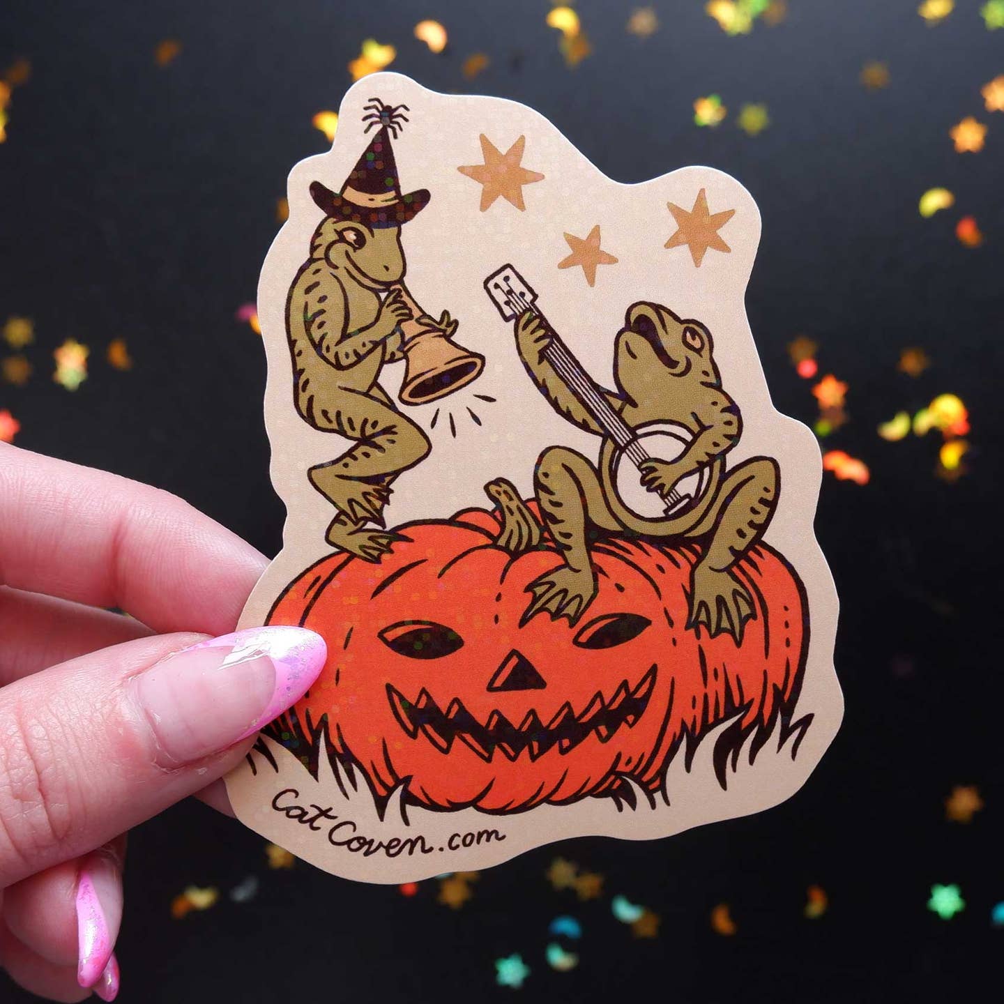 Decorative sticker with tan background and image of two green frogs, playing instruments and sitting on an orange Jack O'Lantern.