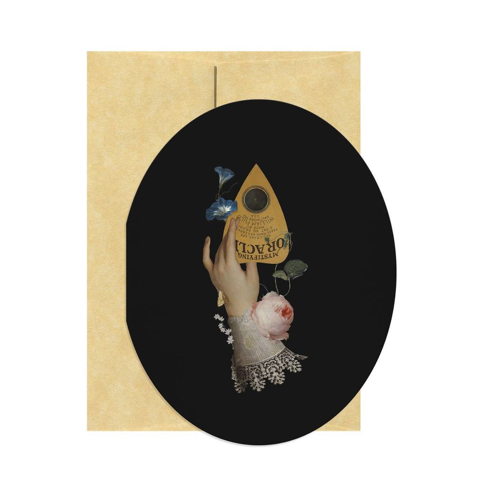 Oval shaped greeting card with black background and image of a hand holding a planchette with flowers and a lace cuff. Tan envelope included.