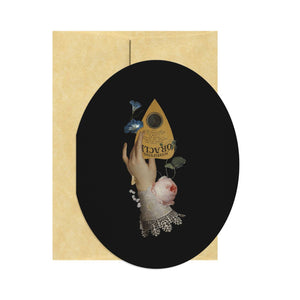 Oval shaped greeting card with black background and image of a hand holding a planchette with flowers and a lace cuff. Tan envelope included.