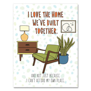 Greeting card with white background and an image of a chair with a table, lam and plant. Orange text says, "I love the home we have built together." and black text says, "And not just beause I can't afford my own place." Envelope included. 