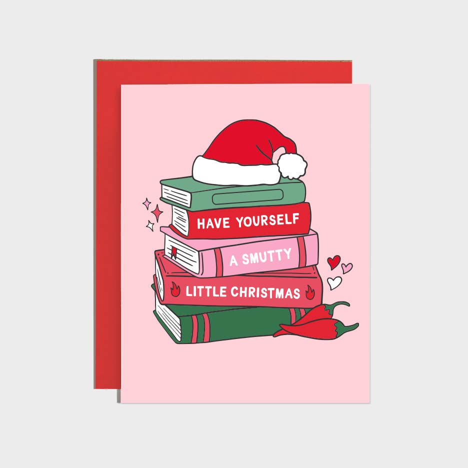 Greeting card with pink background and image of a pile of books and a Santa hat on top and two red hot chili peppers next to them. White text on book binding says, "Have yourself a smutty little Christmas". Red envelope included.