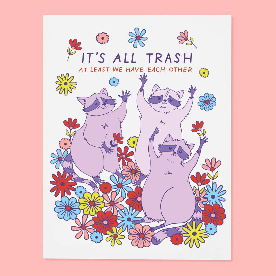 Greeting card with white background and image of three lilac raccoons in a field of flowers. Purple and red text says, "It's all trash at least we have each other". Envelope included.