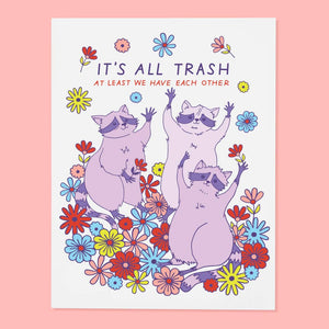Greeting card with white background and image of three lilac raccoons in a field of flowers. Purple and red text says, "It's all trash at least we have each other". Envelope included.