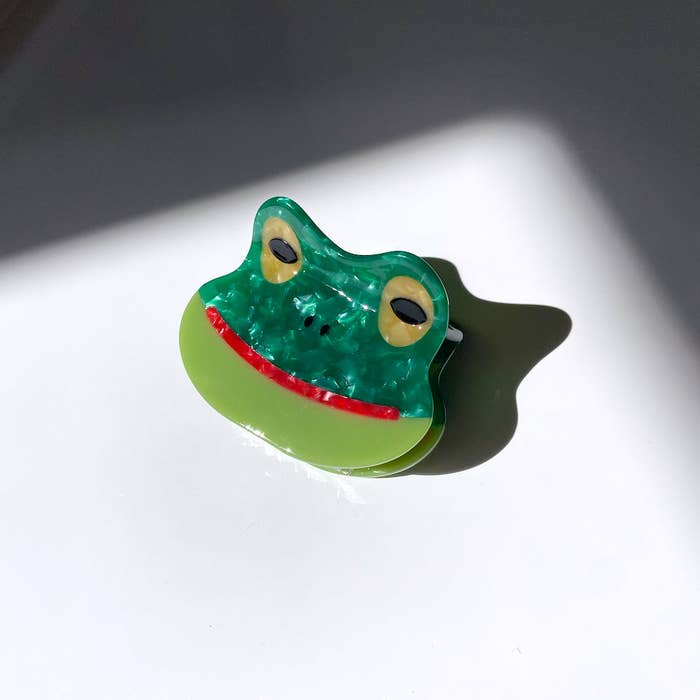 Image of frog face hair clip in dark and light green with yellow eyes and a red mouth. 