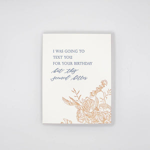 Greeting card with white background and image of flowers in sepia and grey text says, "I was going to text you for your birthday but this seemed better". Grey envelope included.