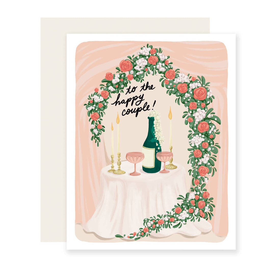 Greeting card with pink background with white border and image of a table  surrounded by flowers in pink, white and green and a bottle of champagne bubbling over  with black text says, "to the happy couple" . White envelope included. 
