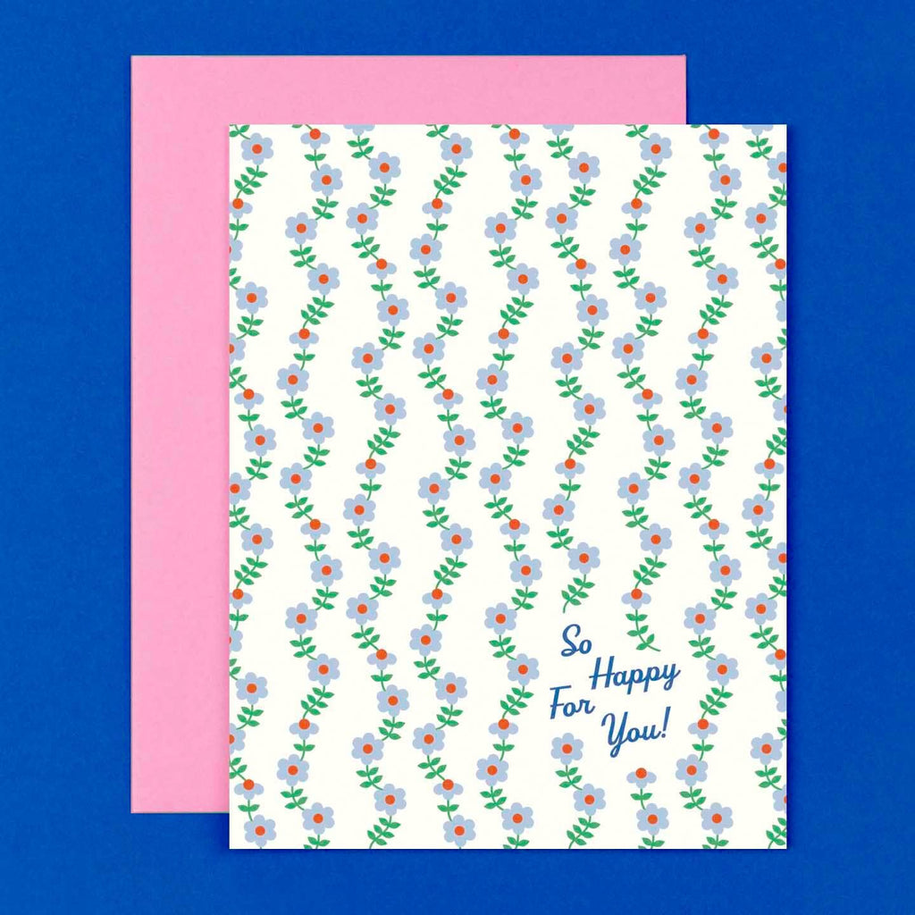 Greeting card with white background and vines of flowers in blue and pink with green stems and blue text says, "So happy for you!". Pink envelope included.