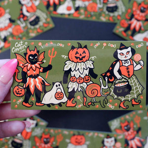 Decorative sticker with olive background and images of three cats in costumes in orange, black and white. 