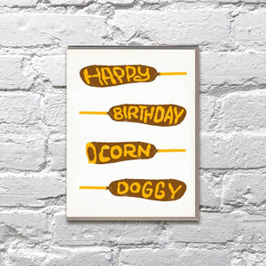 Greeting card with white background and images of four corn dogs in brown with yellow text says, "Happy Birthday Corn Doggy". Envelope included. 
