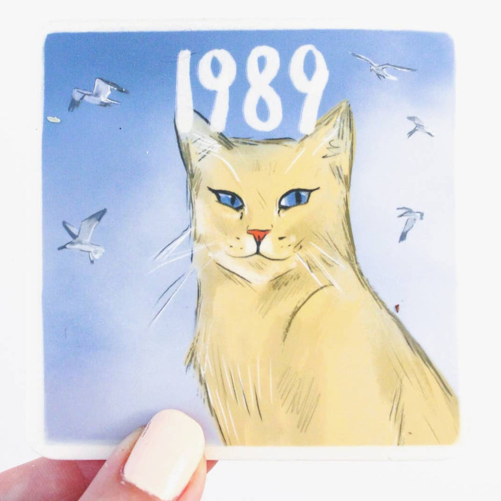 Image of sticker with blue background with white frame and image of tan cat with blue eyes and images of white birds flying in background. White text says, "1989".