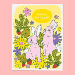 Greeting card with yellow background and forest in green leaves, yellow and purple flowers with two pink bunnies and white text says, "Happy Birthday!". Envelope included. 