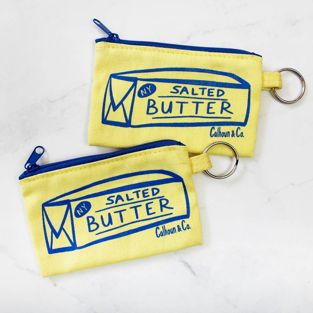 Image of coin purse in light yellow with blue zipper top and metal key chain. Blue printed image of stick of butter. 
