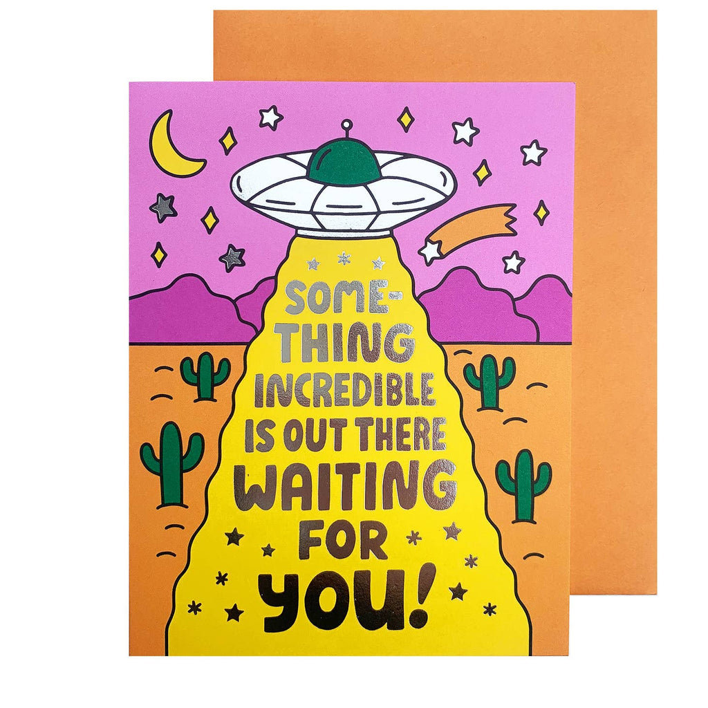 Desert background in pink and orange with white and green spaceship with yellow beam of light. Gold foil text says, "Something incredible is out there waiting for you!". Orange envelope is included. 
