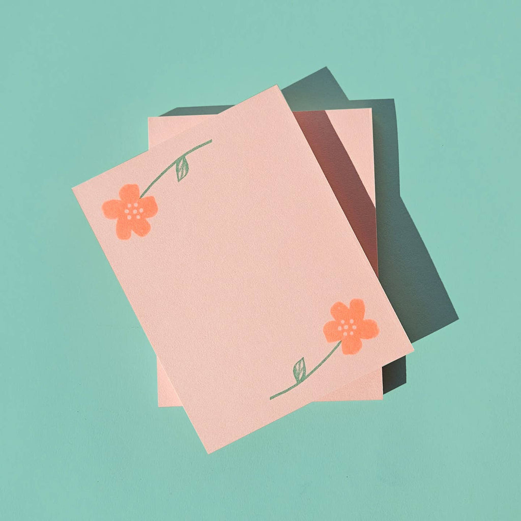 Notepad with pink background with images of peach flowers with green stems, one on top of pad and one at bottom of pad. 