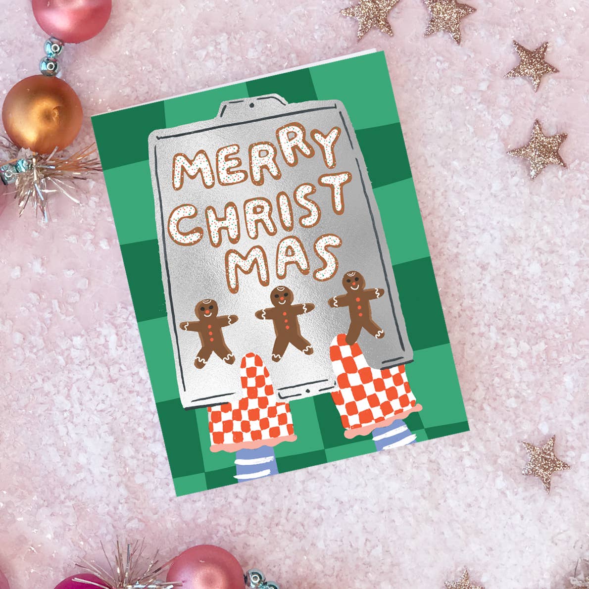 Greeting card with green checkered background and image of a foil sheet pan with images of gingerbread cookies and white text says, "Merry Christmas" being held by red and white checked oven mitts. Envelope included. 