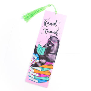Image of bookmark with pink background and image of raccoon wearing pink glasses reading a book while sitting on a stack of books. Black text says, "Read trash".  Green silk tassels at top. 
