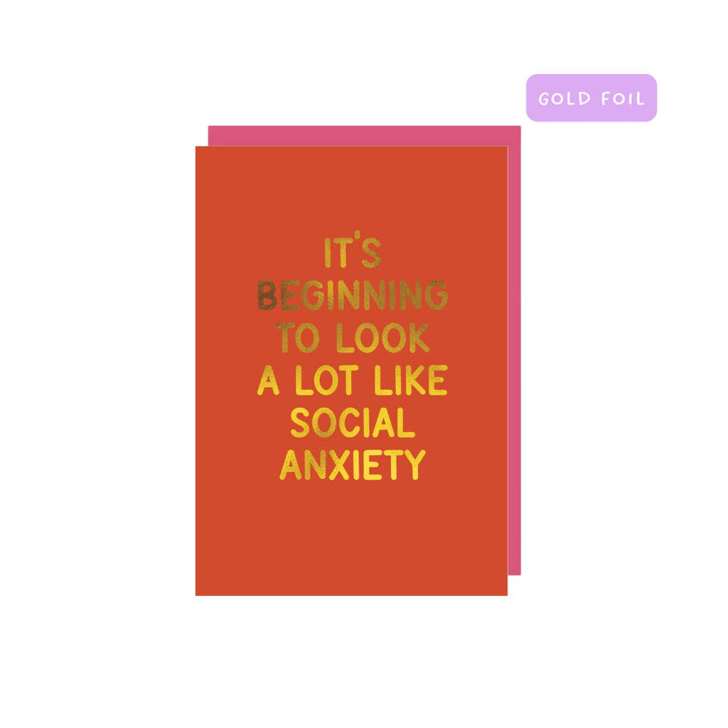 Greeting card with red background and gold foil text says, "It's beginning to look a lot like social anxiety". Pink envelope included. 