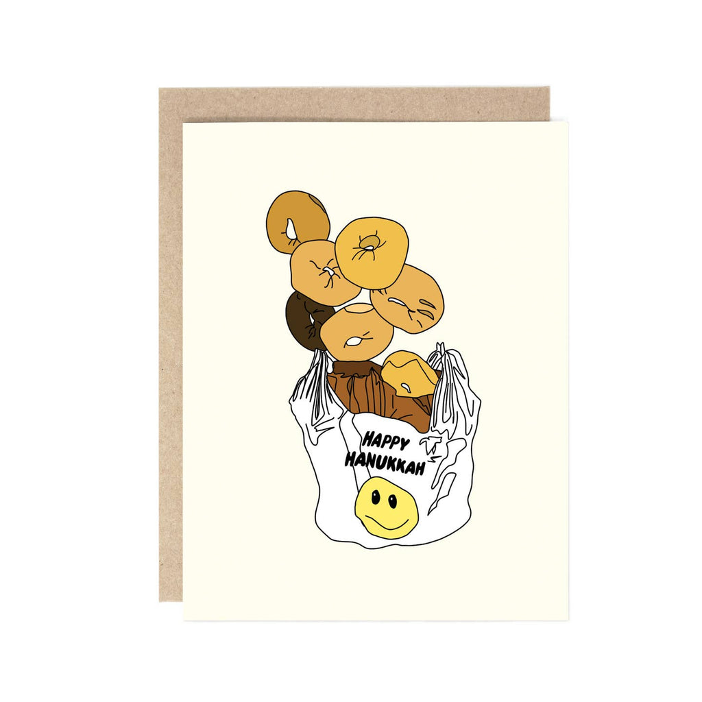 Greeting card with white background and image of a bag of bagels with black text on the white bag says, "Happy Hanukkah" with yellow smiley face.  Kraft envelope included.