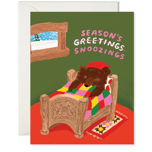 Greeting card with image of a brown bear in a beige bed with a patchwork quilt. Green walls and red floor with a window and a snowy scene outside. Grey envelope included.  Pink and white text says, "Season's Snoozinbs" with "greetings" crossed out.  