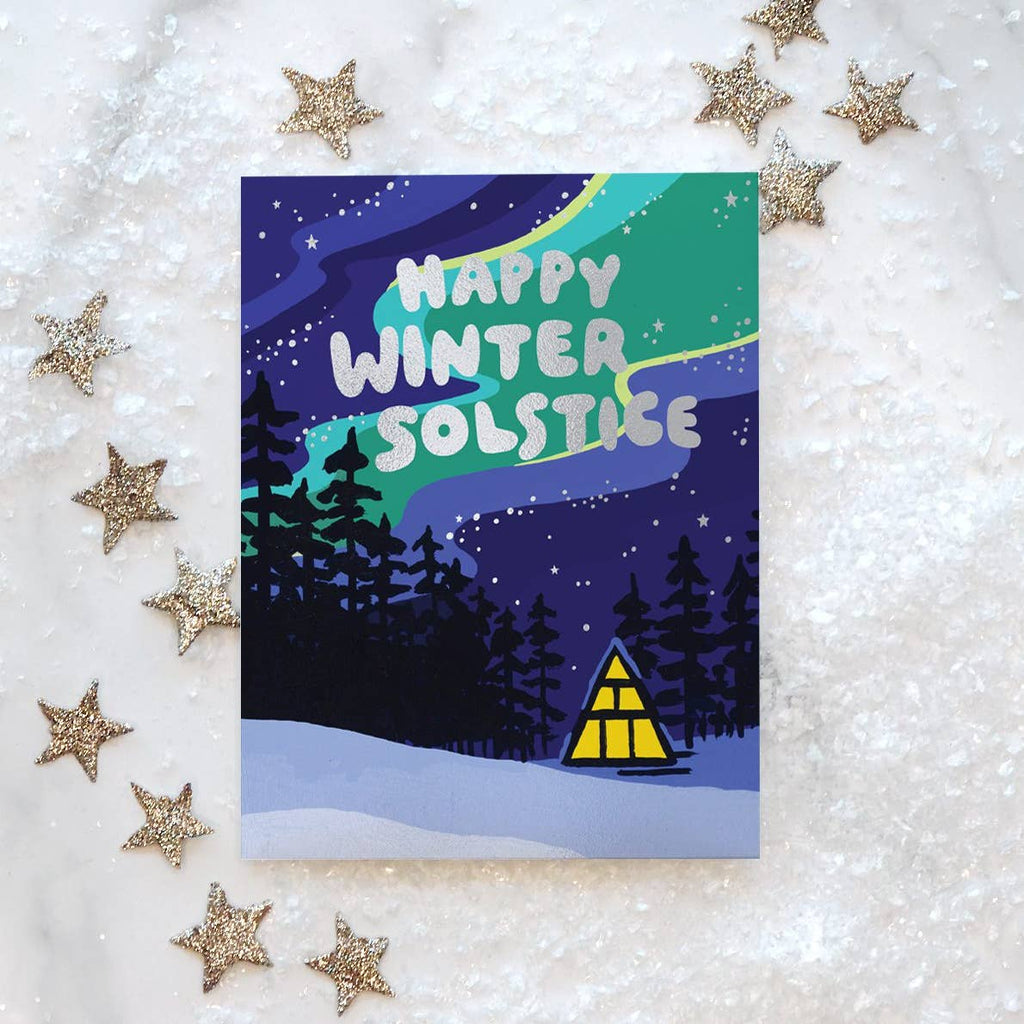 Greeting card with background image of of a night winter sky in blue, green and yellow with foil stars and black trees. Silver foil text says, "Happy Winter Solstice". Envelope included. 