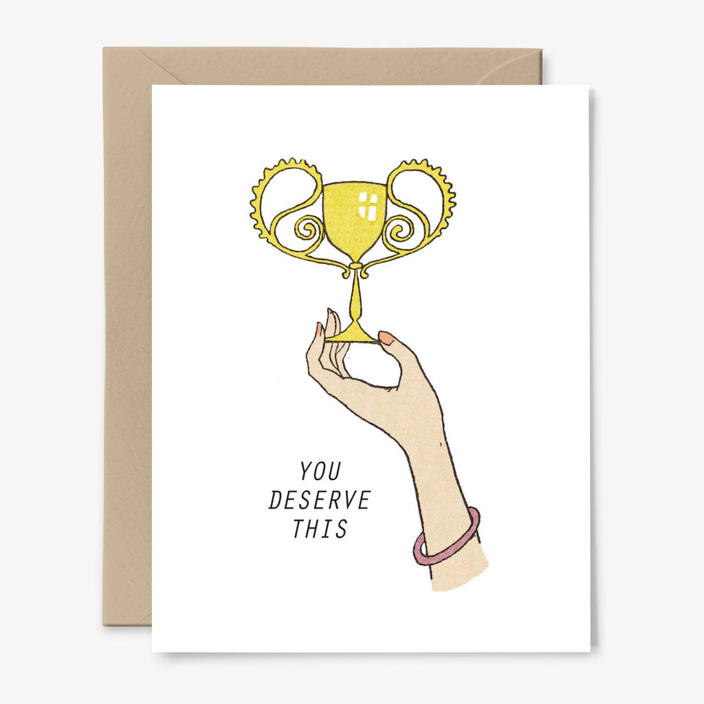 Greeting card with white background and image of a hand holding a trophy cup and black text says, "You deserve this". Envelope included.