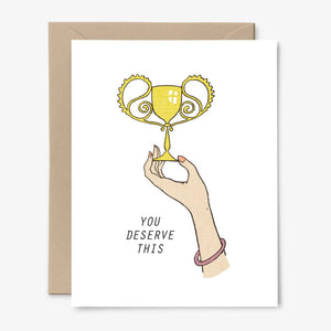 Greeting card with white background and image of a hand holding a trophy cup and black text says, "You deserve this". Envelope included.