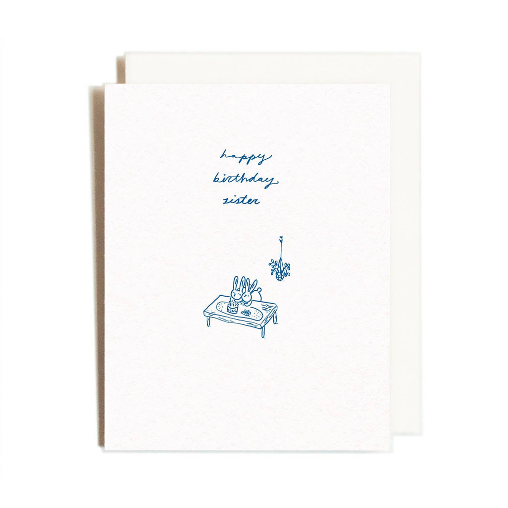Greeting card with cream background and images of two bunnies sitting at a table having cake with blue text says, "Happy birthday sister". Cream envelope included. 