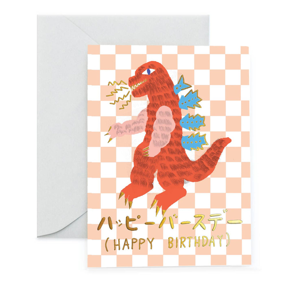 Greeting card with peach and white checkerboard background and image of Godzilla in orange and blue with gold foil text says, "Happy Birthday". Envelope included.