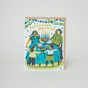 Greeting card with white background and images of a famiy holding hands around a table  of Hanukkah food and a menorah. Yellow text says, "Hanukkah Blessings". Kraft envelope included. 